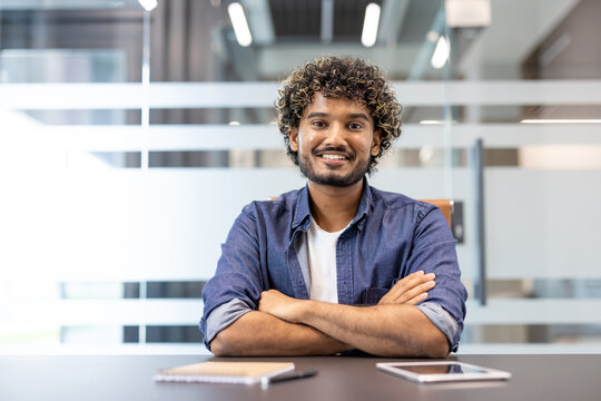 Portrait of an Indian young man in a denim shirt sitting at a desk in the office and smiling at the camera - Powered by Adobe