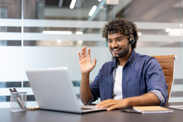 Obraz premium Young Indian man in headset sitting at a desk in the office, talking on a video call on a laptop, greeting and waving
