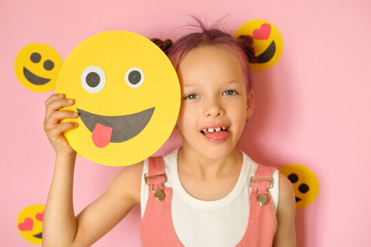 A funny, humorous girl sticks out tongue, holding a grimacing smiley face in hands. World Emoji and Social Media Day.