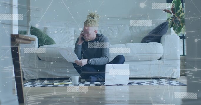African American man talking on smartphone while reviewing papers in living room, with laptop mug