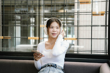 A young woman wearing headphones sits on a sofa, using her smartphone and laptop near a window, enjoying music or working in a relaxed atmosphere.