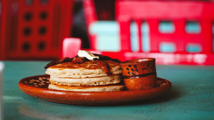 Plato de barro con una torre de hotcakes decorados con tocino y mantequilla, acompañado de una pequeña taza artesanal. La luz cálida resalta la textura esponjosa y el tono dorado del desayuno.
