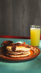 Plato de barro con una torre de hotcakes decorados con tocino y mantequilla, acompañado de una pequeña taza artesanal. La luz cálida resalta la textura esponjosa y el tono dorado del desayuno.