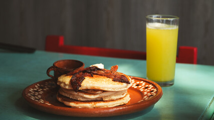 Plato con hotcakes y un vaso de jugo sobre una mesa turquesa. Fondo simple que resalta el desayuno tradicional.