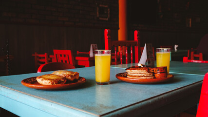 Mesa servida con desayuno completo: hotcakes, jugo, café y miel. Colores vivos y ambiente acogedor...