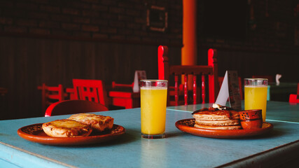 Mesa servida con desayuno completo: hotcakes, jugo, café y miel. Colores vivos y ambiente acogedor que invitan a disfrutar la comida.