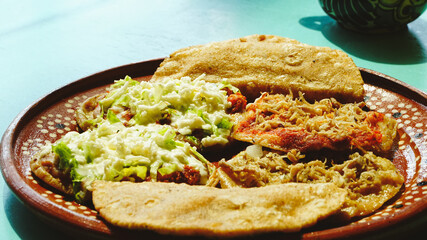 Platillo mexicano con antojitos variados —sopes, tacos dorados y gorditas— servidos en plato de barro sobre una mesa turquesa con luz natural.