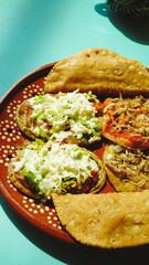 Platillo mexicano con antojitos variados —sopes, tacos dorados y gorditas— servidos en plato de barro sobre una mesa turquesa con luz natural.
