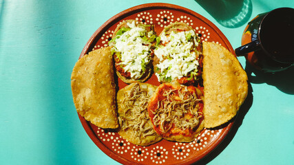 Platillo mexicano con antojitos variados —sopes, tacos dorados y gorditas— servidos en plato de barro sobre una mesa turquesa con luz natural.