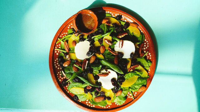 Ensalada fresca servida en un plato de barro, con mezcla de espinacas, aguacate, almendras, pasas y aderezo en una peque&ntilde;a cazuelita. Los tonos verdes y dorados contrastan con la mesa turquesa