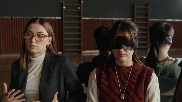 Young confident female school psychologist sitting next to blindfolded teenage girl and explaining or inculcating her how to deal with troubles during hypnosis therapy session