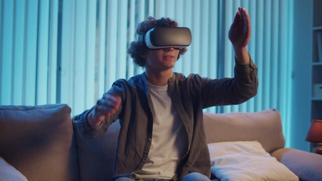 Young Man Sits on Sofa at Night in AR Glasses, Interacting with Virtual Interface. Immersive Experience in Digital World of Tomorrow. Mixed Reality, Future Technology, Mental Escape.