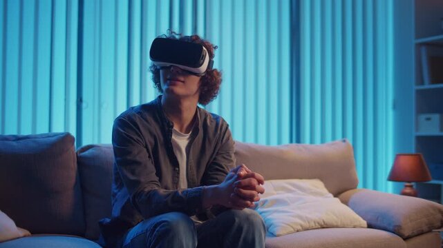 Young Man Sits on Sofa at Night in AR Glasses, Interacting with Virtual Interface. Immersive Experience in Digital World of Tomorrow. Mixed Reality, Future Technology, Mental Escape.