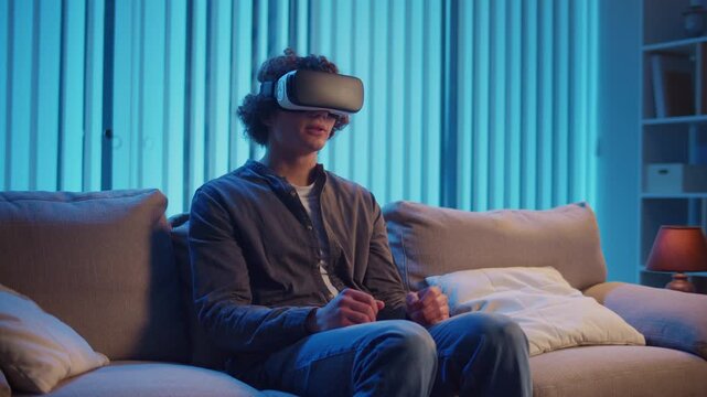 Young Man Sits on Sofa at Night in AR Glasses, Interacting with Virtual Interface. Immersive Experience in Digital World of Tomorrow. Mixed Reality, Future Technology, Mental Escape.