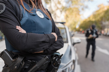 Obraz premium Policewoman standing with arms crossed next to patrol car, other officer working in background