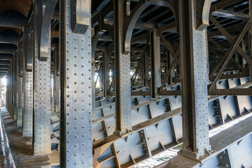 Underneath a historic bridge, its intricate riveted steel structure unfolds, revealing repetitive columns, arched elements, and crossbeams, with sunlight accentuating industrial details above water.
