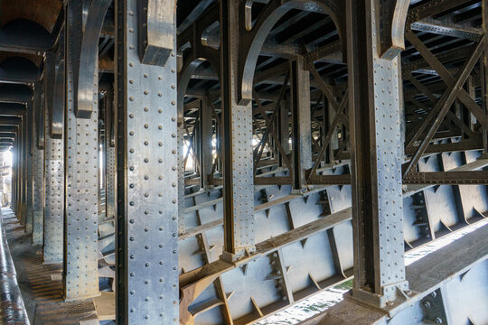 Underneath a historic bridge, its intricate riveted steel structure unfolds, revealing repetitive columns, arched elements, and crossbeams, with sunlight accentuating industrial details above water.