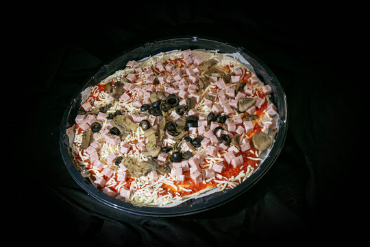 Uncooked pizza topped with diced ham, sliced mushrooms and black olives in a takeaway tray
