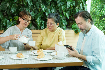Asian senior woman smiles as a Caucasian woman pours tea at a cafe table with another Caucasian friend, all enjoying pastries and sharing a joyful and relaxed retirement afternoon.