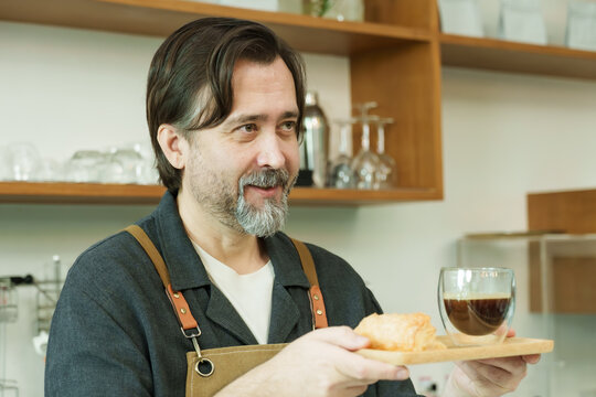 Senior Caucasian male barista wearing brown apron smiling while holding tray with croissant and coffee drink ready to serve customer inside small cafe setting with equipment., Concept small business