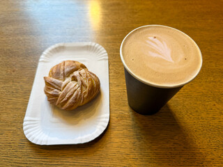 Coffee latte art on wood table with a bun with lemon in cafe close-up. Cappuccino with a pattern in a paper cup and bun on a wooden table in a coffee shop top view. The concept of food and drinks.