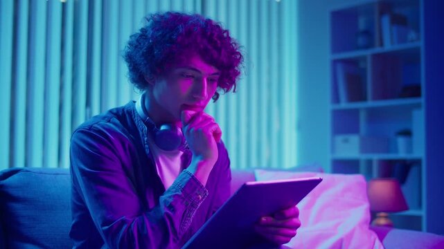 Young Man Sits on Sofa with Tablet in Hands, Working with Concentration. Calm and Efficient Evening in a Flexible Tech-Driven Workflow. Focus Mode, Digital Productivity, Self-Management.