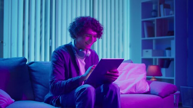 Young Man Sits on Sofa with Tablet in Hands, Working with Concentration. Calm and Efficient Evening in a Flexible Tech-Driven Workflow. Focus Mode, Digital Productivity, Self-Management.