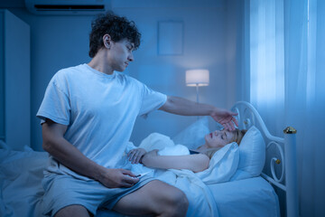 Obraz premium A man sits beside a sleeping woman suffering from a cold in a dimly lit bedroom