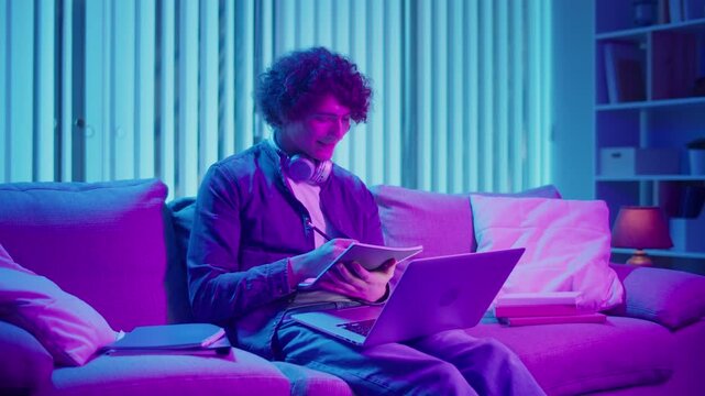 Young Man Studies at Night on Sofa in Neon Light, Holding Notebook and Laptop. Gen Z Study Routine with Calm Productivity and Mood Lighting. Remote Learning, Digital Journaling, Aesthetic Focus.