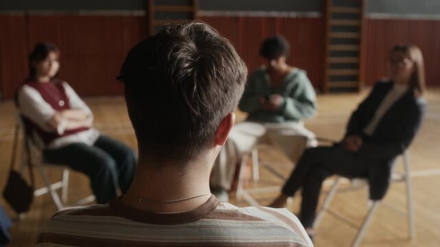 Rear view of youthful guy wearing striped pullover sitting in front oif his schoolmates and female counselor at session and speaking about his psychological problems