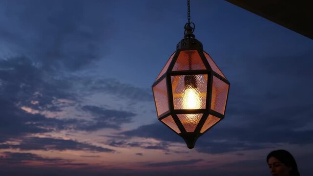 A lit geometric lantern with textured panels hangs from a chain against a cloudy twilight sky showing blue and purple hues A persons blurred profile is visible in the lower right