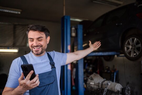 Happy mechanic showing a lifted car while using a smartphone in - Powered by Adobe