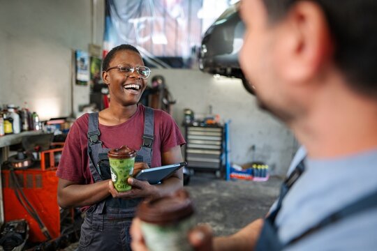 Cheerful african american female mechanic laughing with colleague during coffee break at auto repair shop