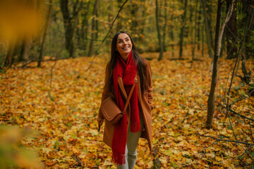 Smiling young woman walking through autumn forest, wearing brown coat and red scarf, enjoying peaceful nature walk surrounded by colorful yellow leaves and warm seasonal light.
