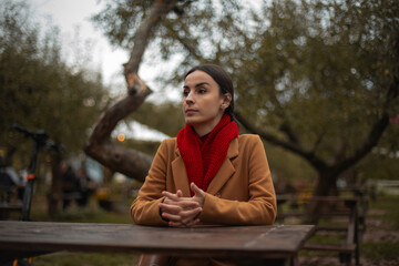 Thoughtful young woman in brown coat and red scarf sitting alone at wooden table in autumn park, looking away pensively, calm mood, outdoor portrait in soft evening light.