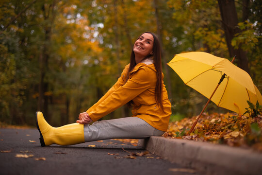 Smiling young woman in yellow raincoat and boots sitting on autumn road with yellow umbrella, enjoying rainy weather and nature, surrounded by colorful fall leaves.