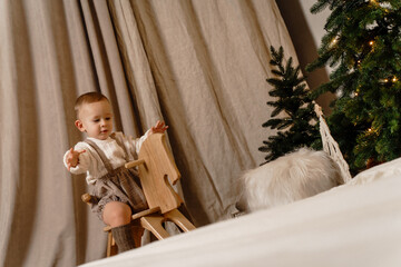 Little boy in casual overalls and socks rides a rocking horse near a Christmas tree