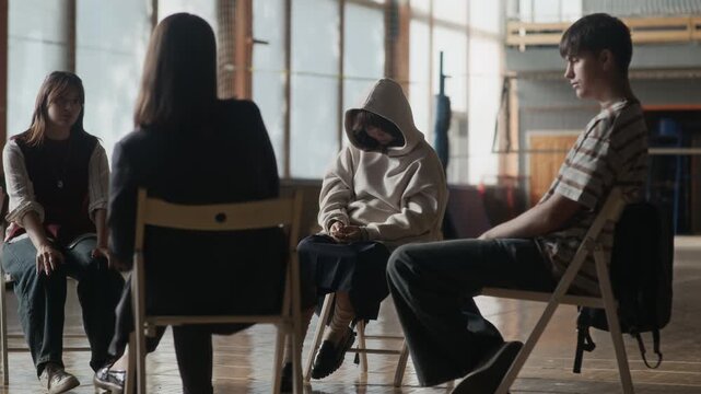 Troubled Asian teenager putting hood on head while sitting in front of school psychologist and two schoolmates discussing ways of solving problems of youth