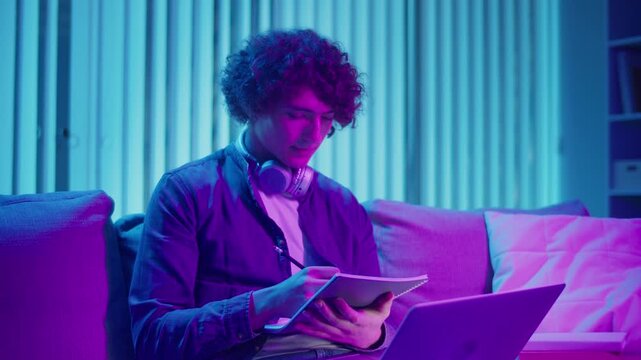 Young Man Studies at Night on Sofa in Neon Light, Holding Notebook and Laptop. Gen Z Study Routine with Calm Productivity and Mood Lighting.