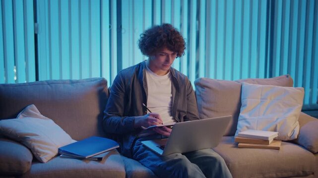 Young Man Studies at Night on Sofa in Neon Light, Holding Notebook and Laptop. Gen Z Study Routine with Calm Productivity and Mood Lighting. Remote Learning, Digital Journaling, Aesthetic Focus.
