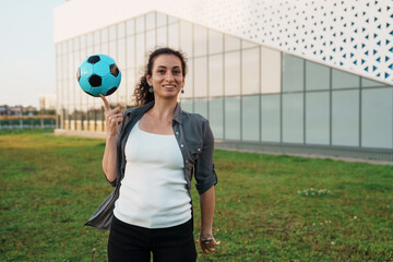 A woman spins a soccer ball on her finger, standing confidently in front of a modern building,...