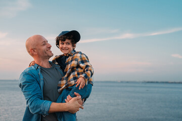 A man holds a young boy joyfully by the sea with a cap on his head, both smiling broadly,...