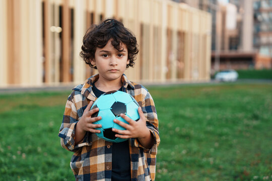 A young boy with curly hair holds a blue soccer ball in a grassy field, framed by modern wooden architecture, conveying a sense of playful curiosity.