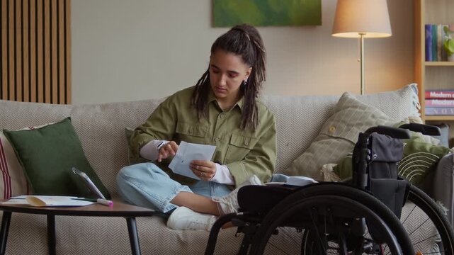 Full shot of young Caucasian girl with dreadlocks in casualwear sitting on couch in cozy living room near wheelchair, opening utility bill letter and analyzing monthly expenses