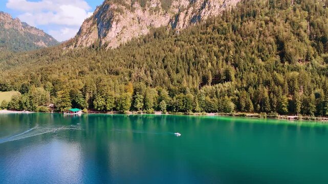 Turquoise lake with a boat. Pine tree woods grow on the rocks at the lakefront. Beautiful buildings and Neuschwanstein Castle at backdrop. Bavaria, Germany.