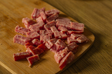 Small pieces of diced sausage are arranged on a wooden cutting board. The pink meat is ready for cooking, placed on a wooden kitchen surface during meal preparation.
