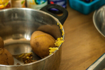 A stainless steel pot with unpeeled boiled potatoes sits on a wooden countertop. Potato peels cling to the rim, hinting at meal prep in progress in a casual kitchen setting.