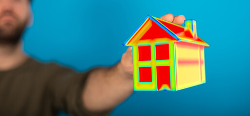A person holds a miniature house model with a heat map visualization. The background is plain blue,...