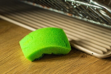 A used green sponge rests on a wooden kitchen counter beside a drying rack. The sponge shows signs of wear, indicating frequent use for dishwashing or cleaning in a domestic setting.