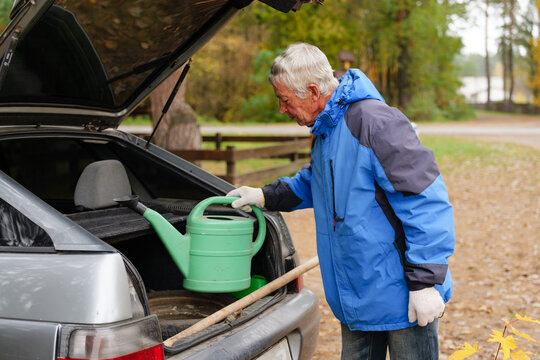 An elderly man in a blue jacket prepares a green watering can while standing near his car in a serene forest setting in autumn
